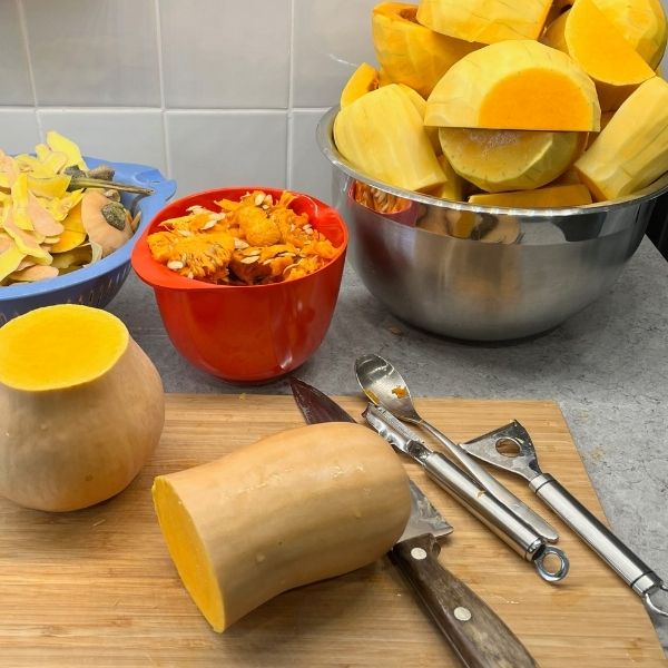 Butternut squash being prepared in a kitchen: peeled and halved squash on a cutting board, a bowl of peels, a bowl of seeds, and kitchen utensils on a countertop.