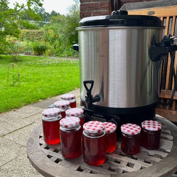 Homemade jars of pink rhubarb juice cooling on a wooden table next to a stainless steel water bath canner, set on a patio with a garden in the background.