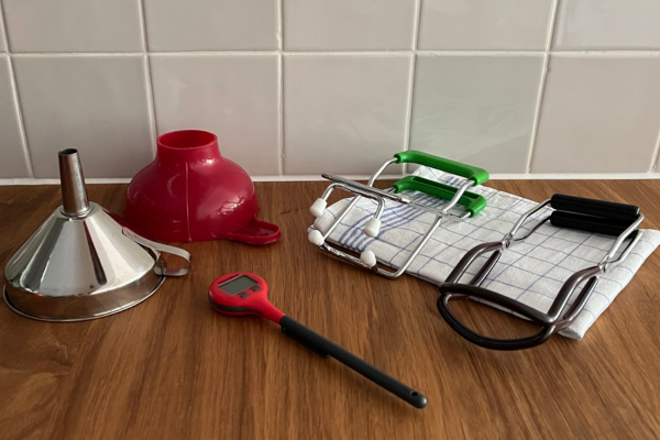 Canning supplies on a wooden surface: funnels, jar lifters, dish towel, thermometer
