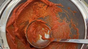 Pureed bell peppers and tomatoes being pushed through a mesh sieve to remove seeds